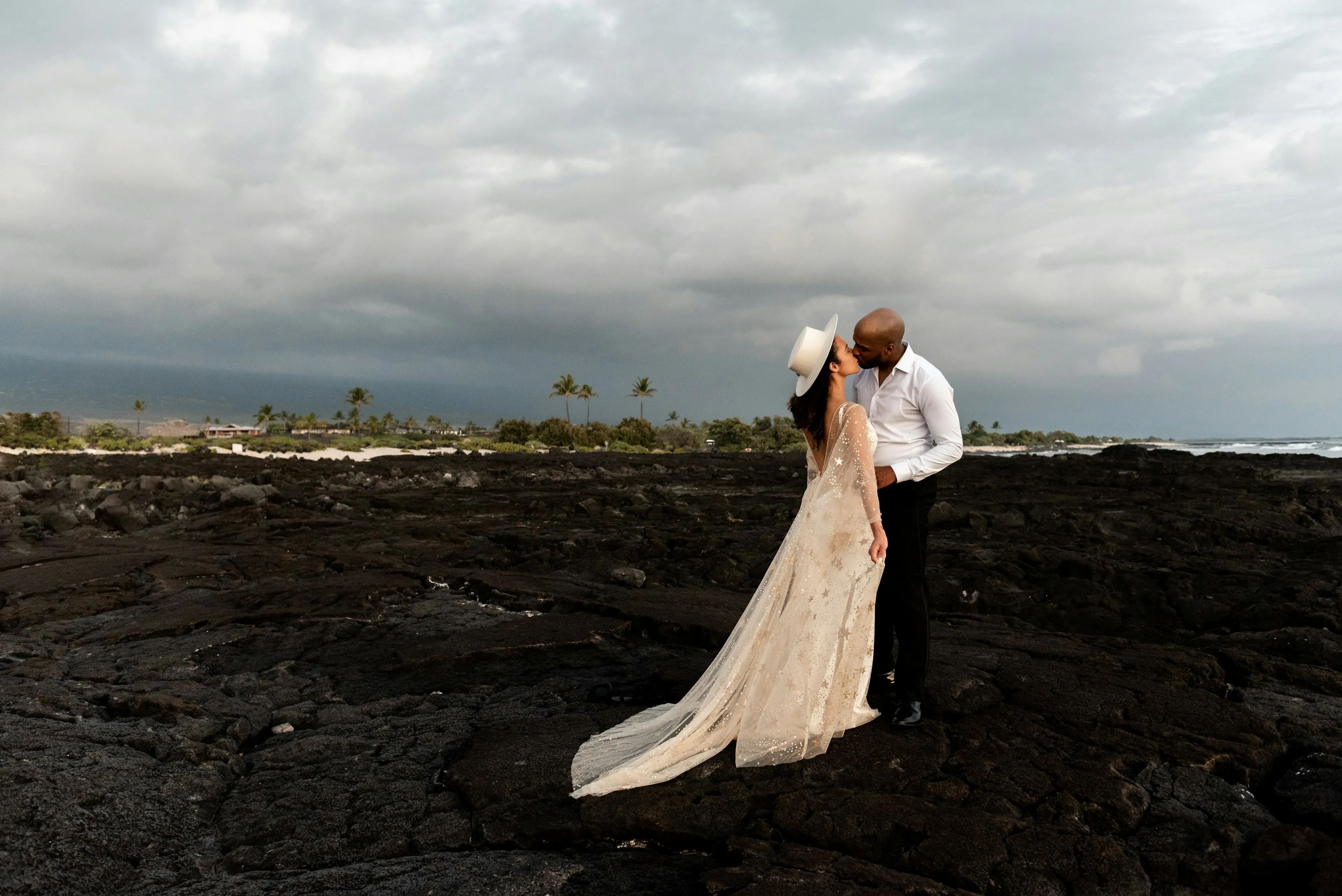 A bride and groom kissing on black lava rock under a storm sky